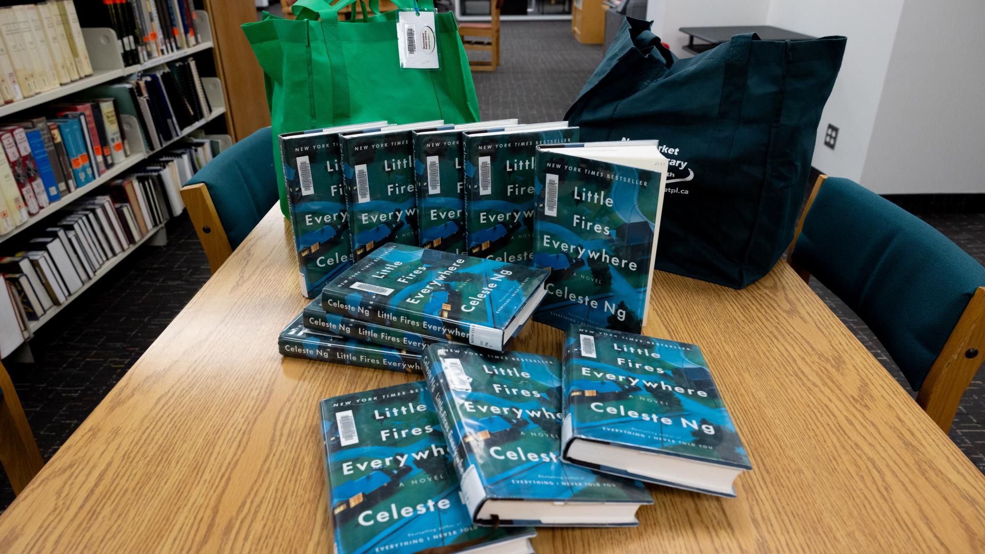 Stack of books titled "Little Fires Everywhere" on a library table, with bags nearby.