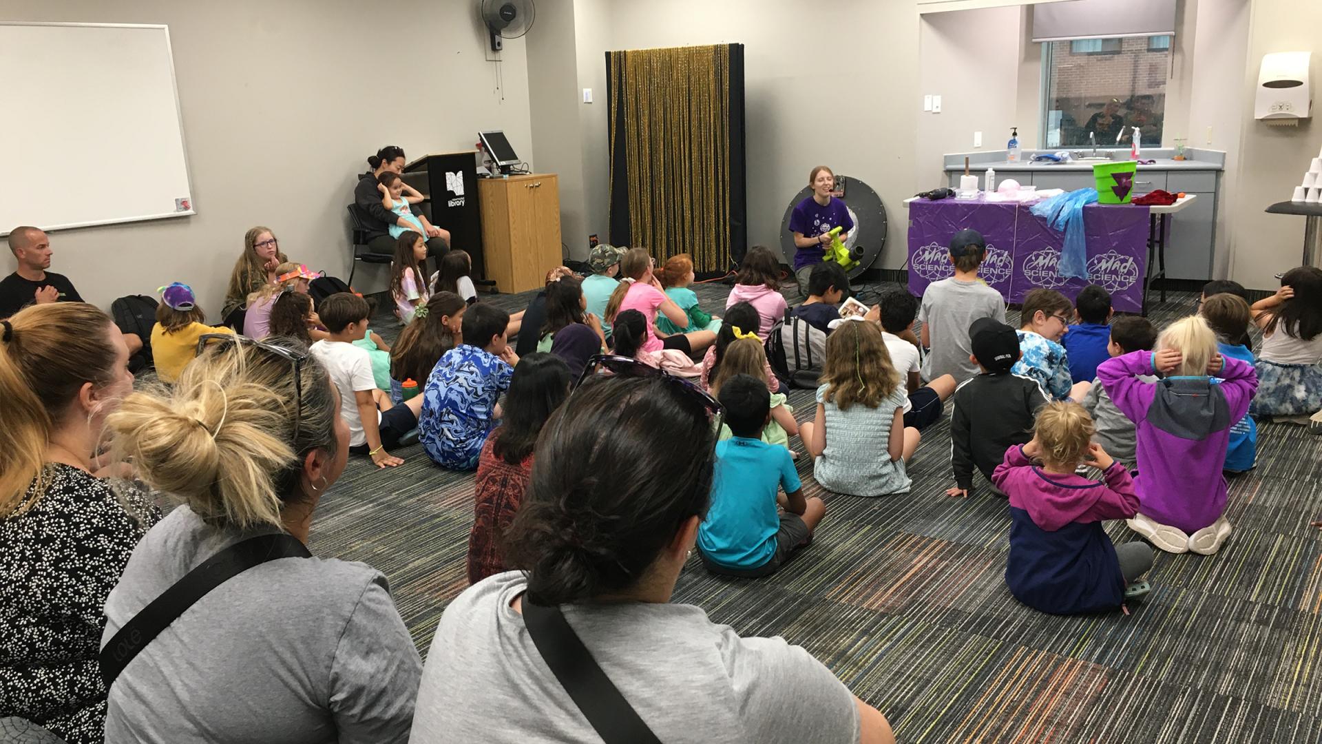Children sitting on a library floor, listening to a storyteller at the front.