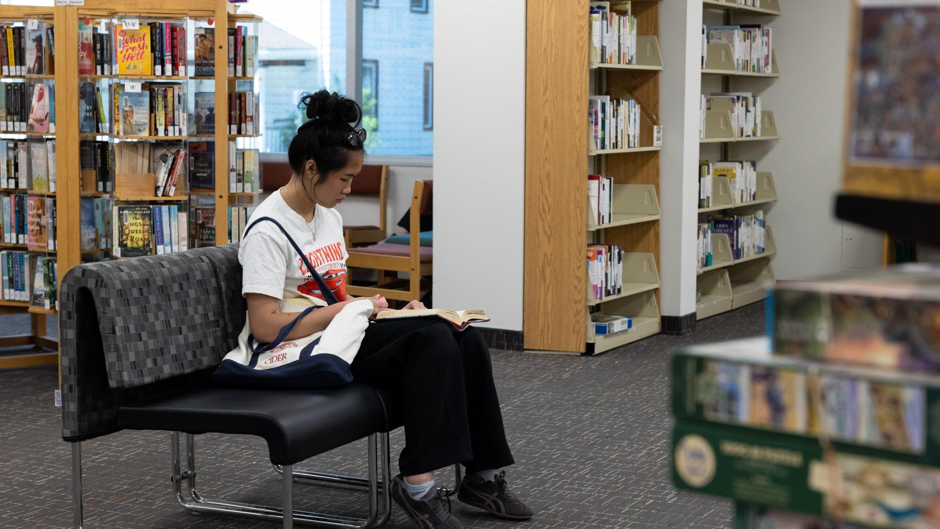 Woman reading on a bench in a library. Bookshelves surround her.