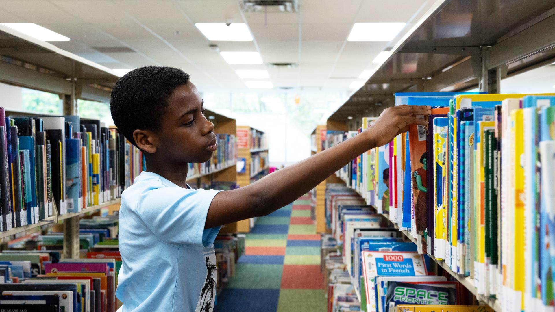 A child in a library reaches for a book on a colorful shelf.