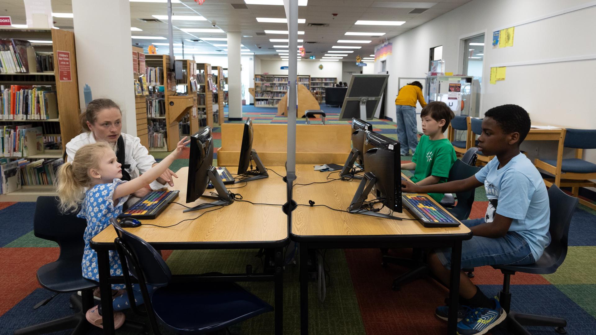 Children sitting at computer stations in a library, engaged in activities.