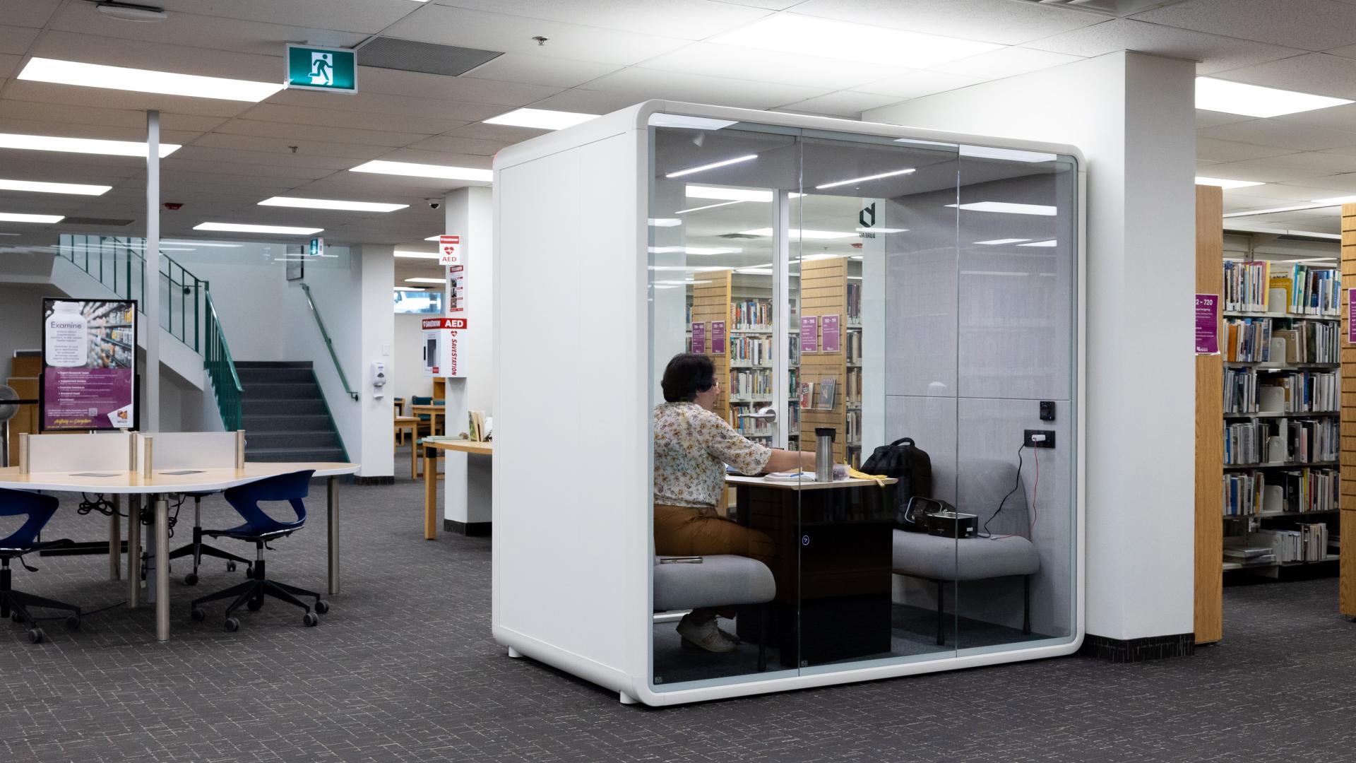 Library study pod with person inside, surrounded by bookshelves and tables.