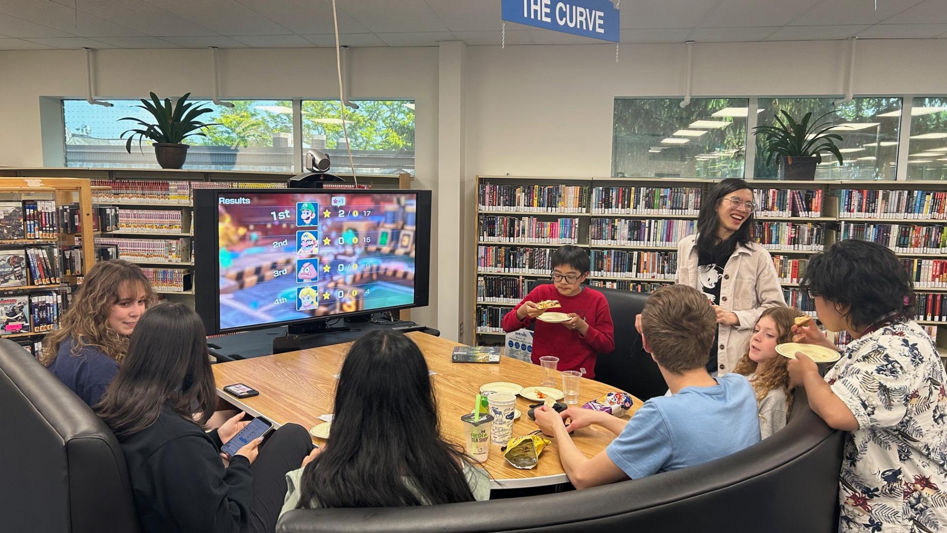 Students gathered around a table in a library, watching a large screen.