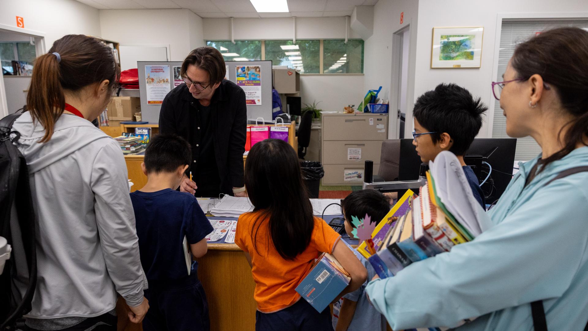 People at a library desk, children holding books, talking to a librarian.