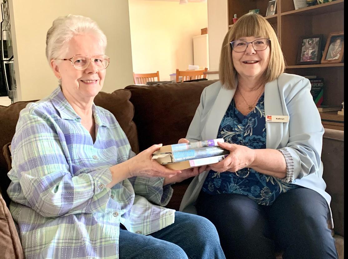 Two women sitting on a sofa, exchanging books, smiling warmly.